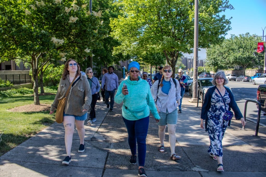 A group of people walk together outdoors on a sunny day, surrounded by green trees and parked cars.