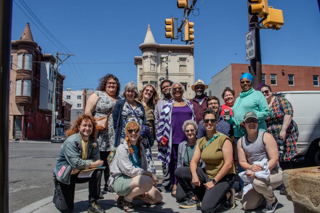 A group of people poses and smiles on a sunny city street corner with buildings in the background.