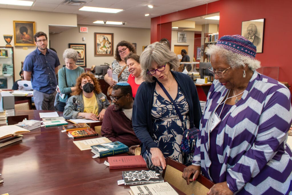 A group of people examine books and papers on a long table in a library or archive room.