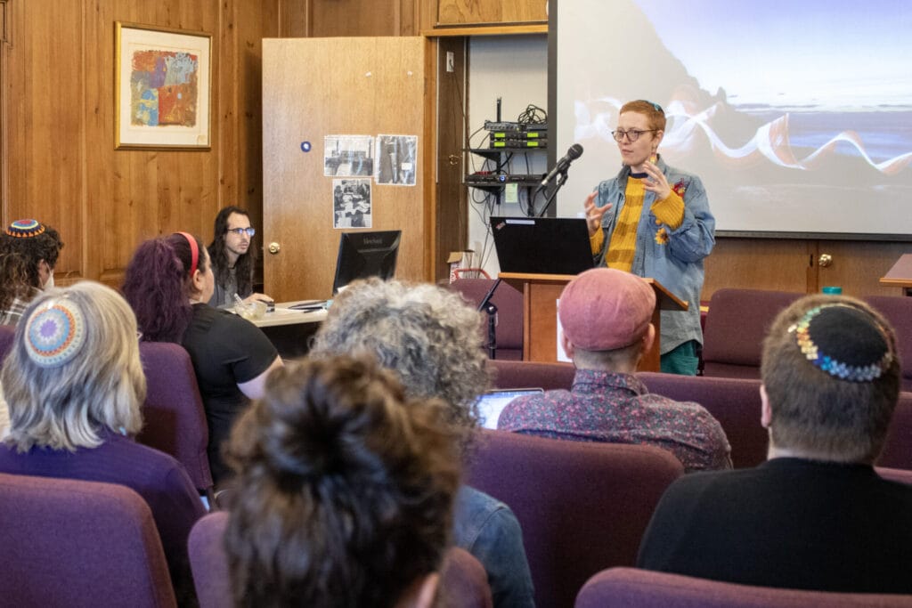 A person speaks at a podium to an audience in a wood-paneled room with a projector screen.