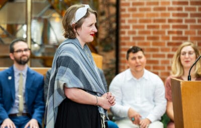 A woman with a headband and shawl stands speaking, with three people seated behind her indoors.