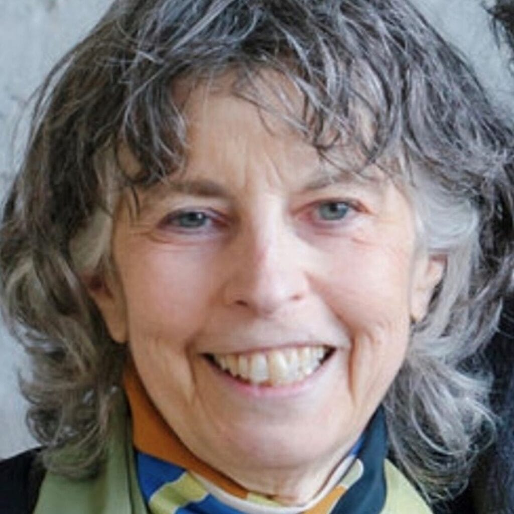 Smiling older woman with gray curly hair and a colorful scarf, standing in front of a neutral background.