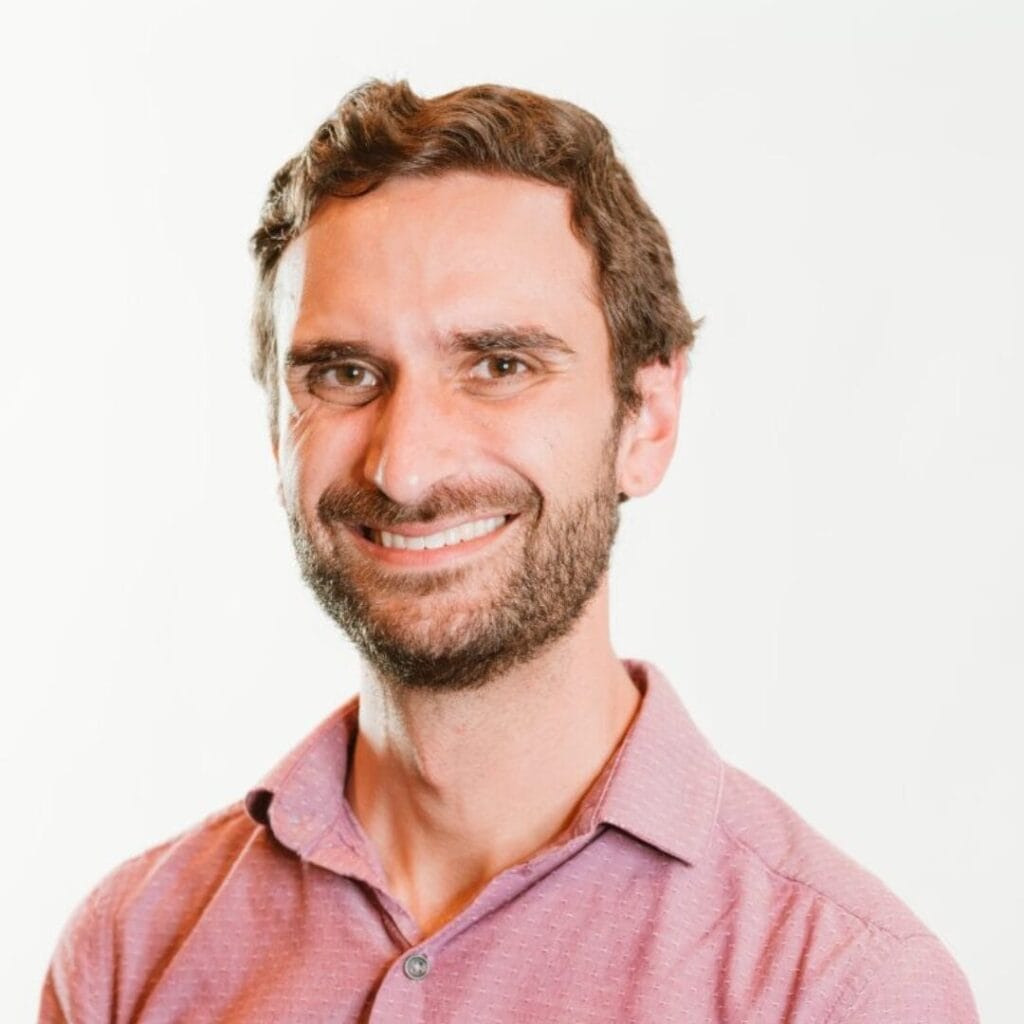 A man with brown hair and beard smiles, wearing a light pink collared shirt, on a white background.