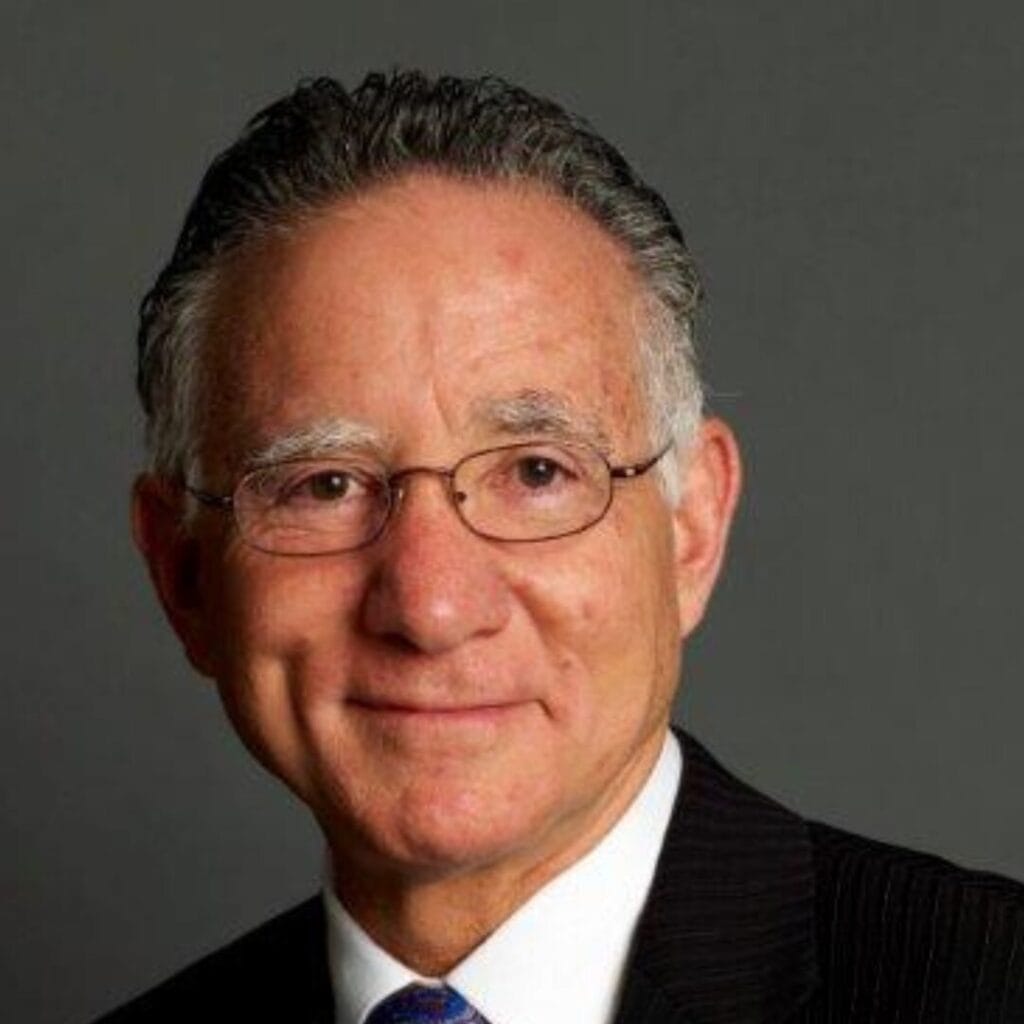 An older man with glasses in a suit and tie, smiling in front of a plain gray background.