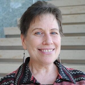 Smiling woman with short hair, wearing patterned shirt and earrings, standing in front of wooden steps.