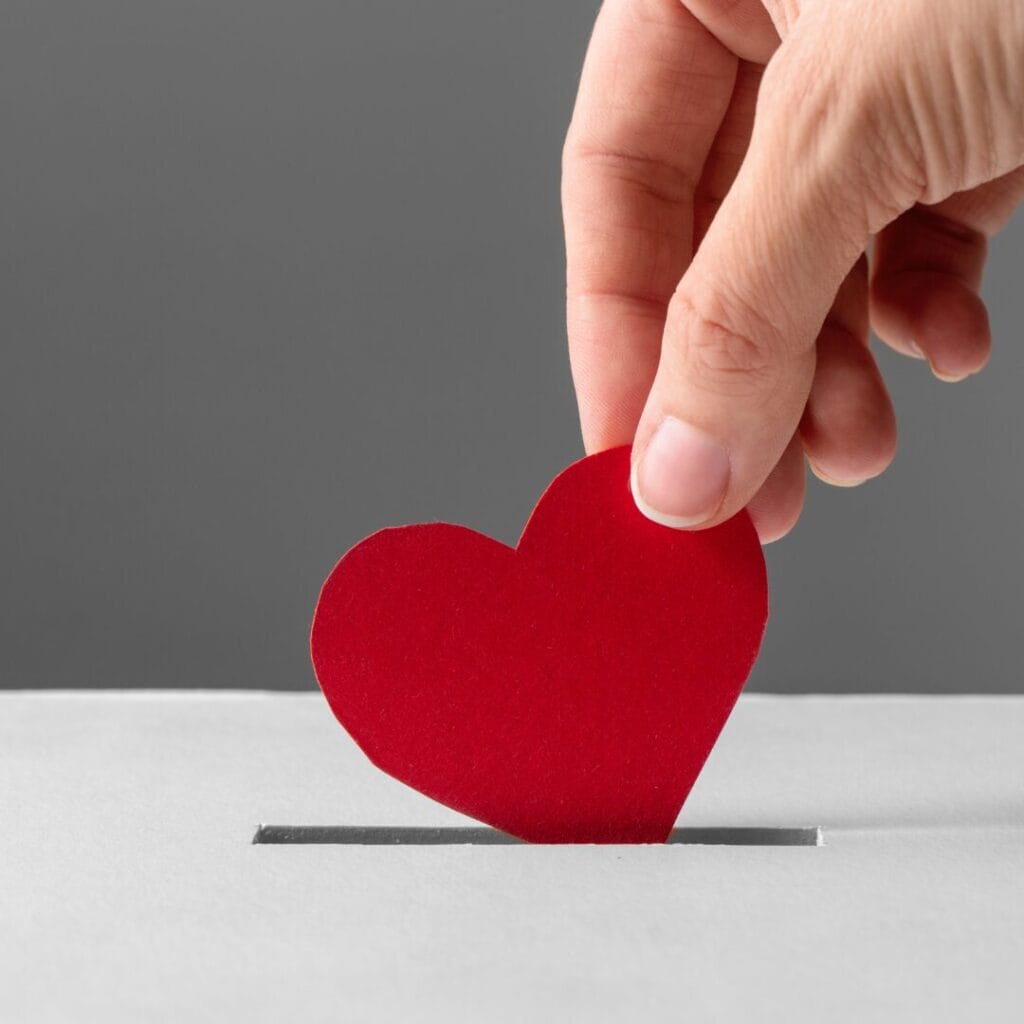 A hand places a red paper heart into a slot, resembling a donation or ballot box, on a gray background.