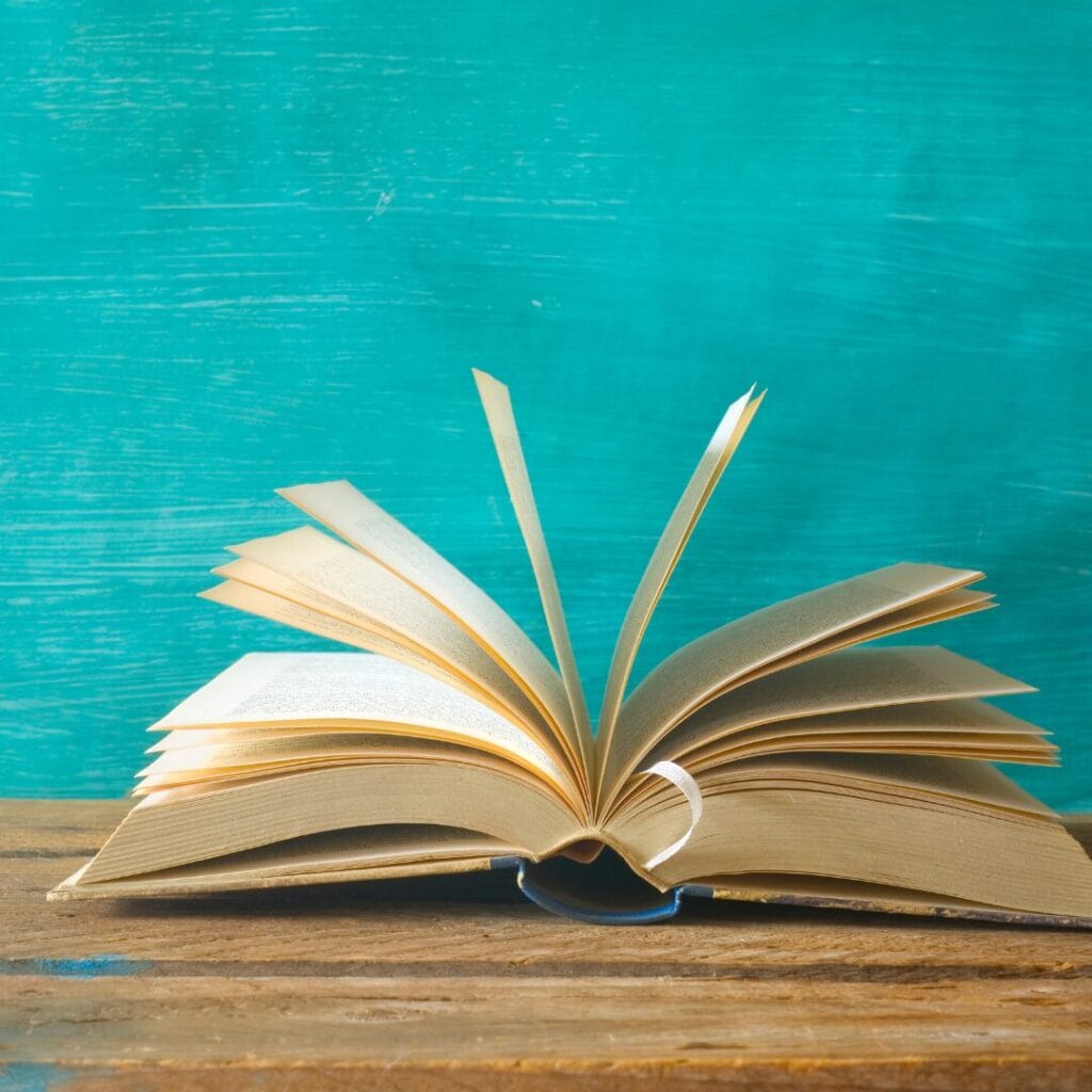 An open book with pages flipping, set on a wooden table against a teal background.