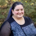 A woman with long dark hair smiles outdoors, wearing a black and gray top; greenery is in the background.