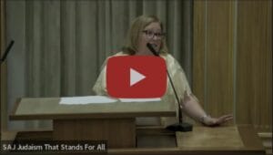 A woman speaks at a podium in a synagogue with papers and a microphone in front of her.