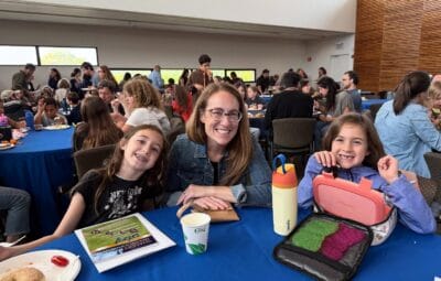 A woman and two girls smile at a table during a busy indoor event with food and drinks.
