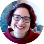 Woman with short dark hair, glasses, and a red top smiling in a brightly lit room.