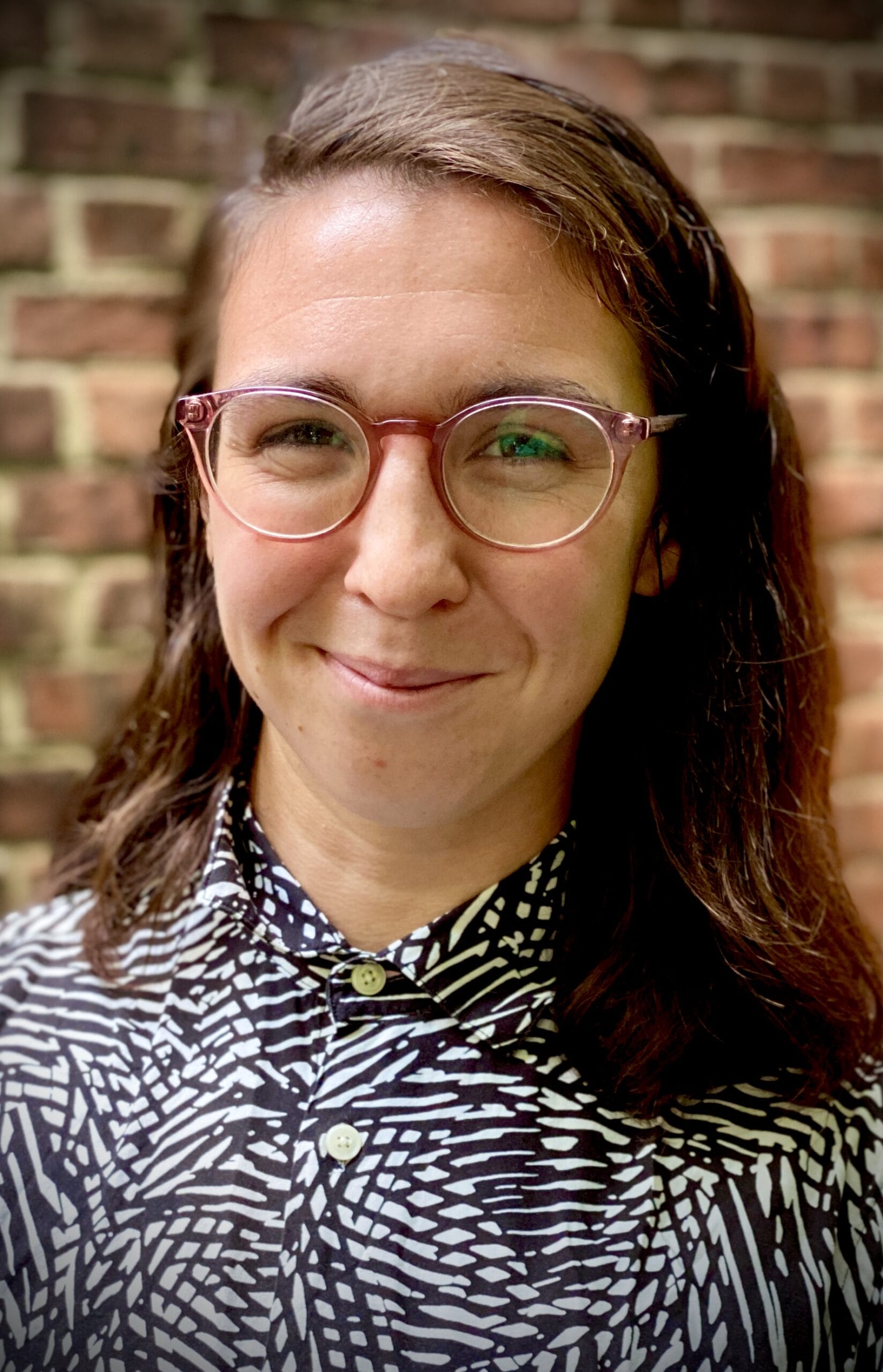 Smiling person with glasses and patterned shirt standing in front of a brick wall.