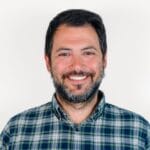 A man with a beard and mustache, wearing a plaid shirt, smiling in front of a plain white background.