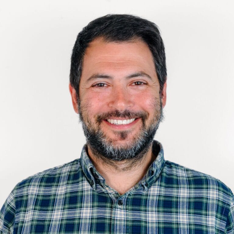 A man with a beard and mustache, wearing a plaid shirt, smiling in front of a plain white background.