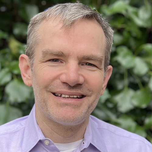 Smiling man with short gray hair wearing a light purple shirt, standing in front of green leafy plants.