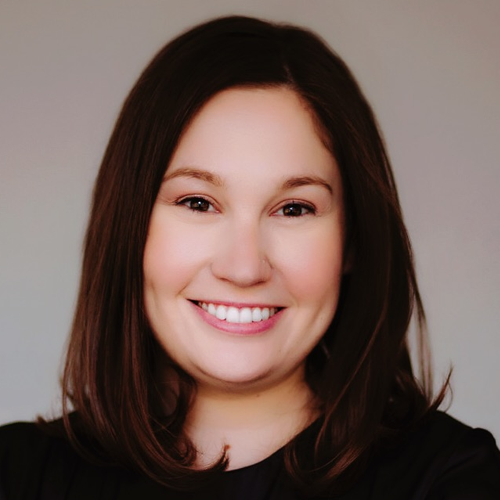 A woman with straight brown hair smiling, wearing a black top, against a neutral background.