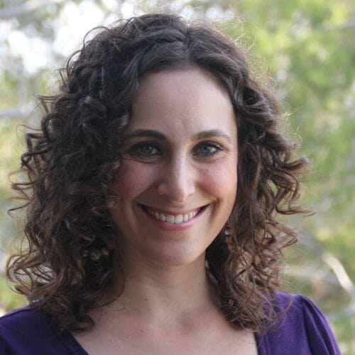 Smiling woman with curly brown hair standing outdoors in front of green trees, wearing a purple top.