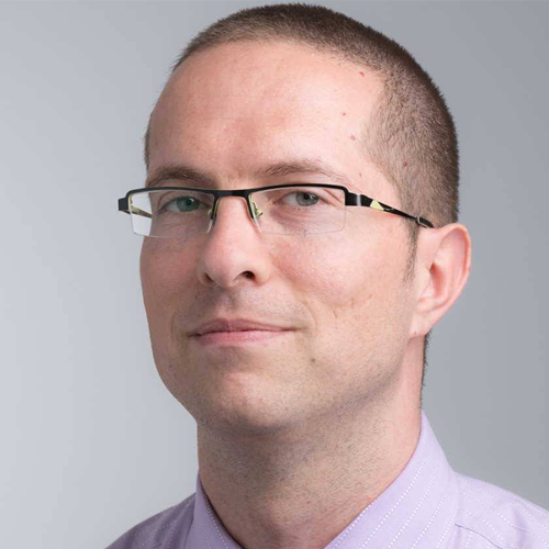 Man with short hair, glasses, and a light purple shirt, smiling slightly against a gray background.