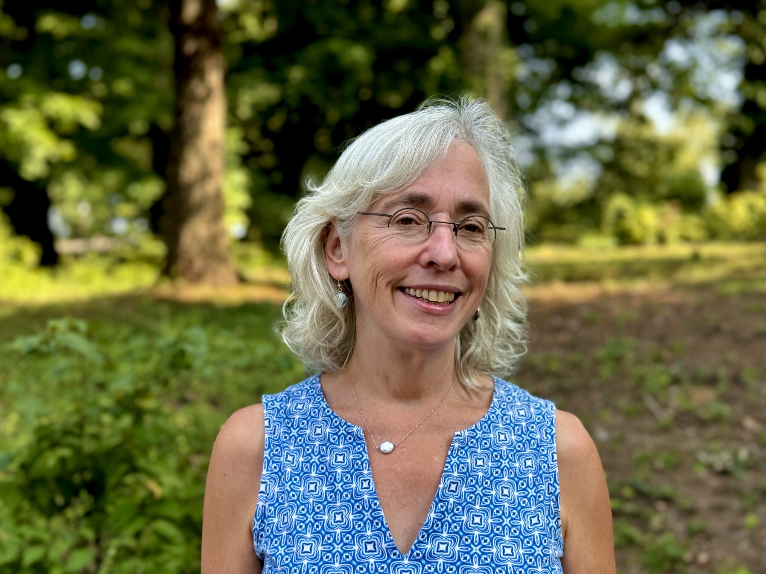 Woman with gray hair and glasses smiling outdoors, wearing a blue patterned sleeveless top with greenery behind her.