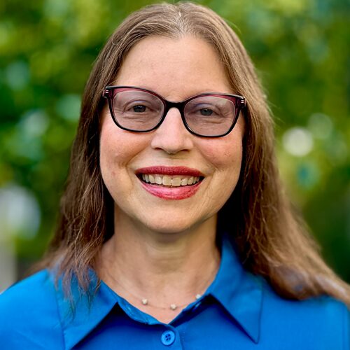 Woman with long brown hair and glasses smiling, wearing a blue shirt, with a green blurred background.