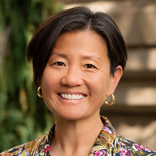 Smiling woman with short dark hair, gold hoop earrings, and a floral top, outdoors with greenery in background.