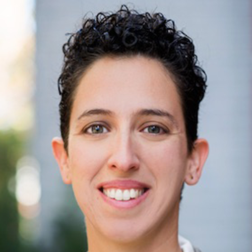 Person with short, curly dark hair smiling outdoors with a blurred background.