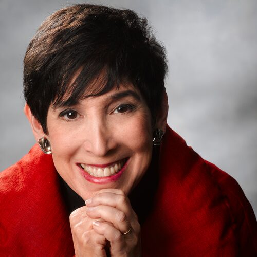 Smiling woman with short dark hair, wearing a red jacket and silver earrings, hands clasped under chin.