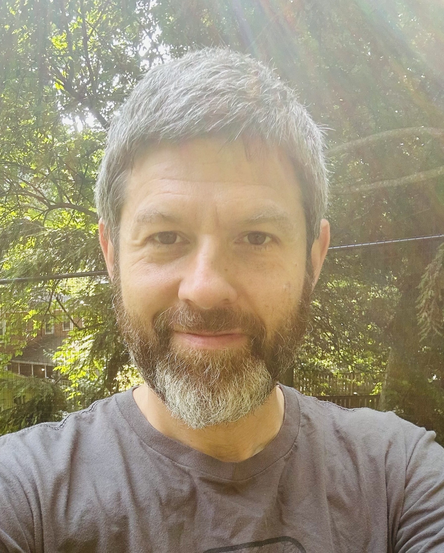 Smiling man with gray hair and beard stands outside with sunlight filtering through trees behind him.
