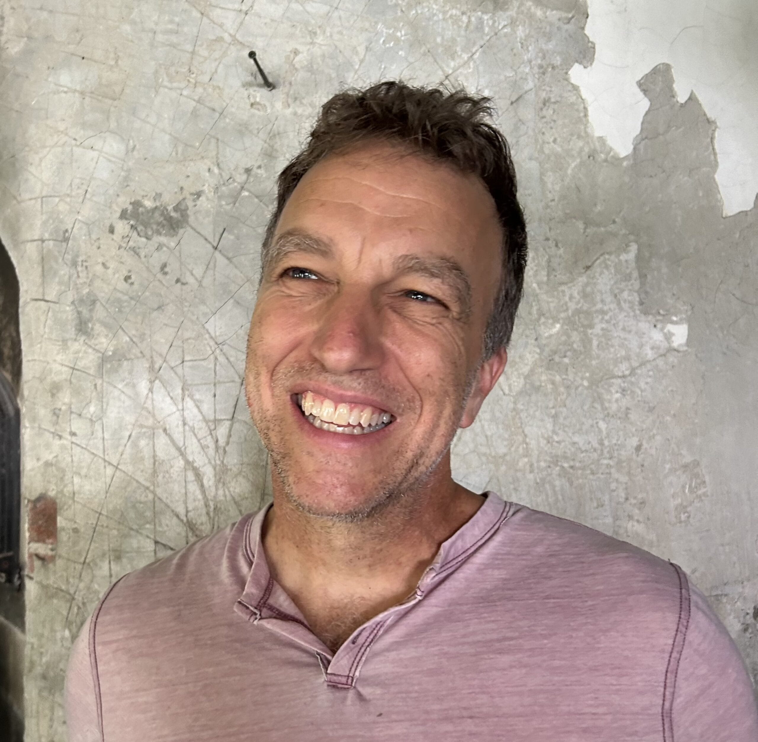 Man smiling in front of a weathered, textured wall while wearing a light pink shirt.
