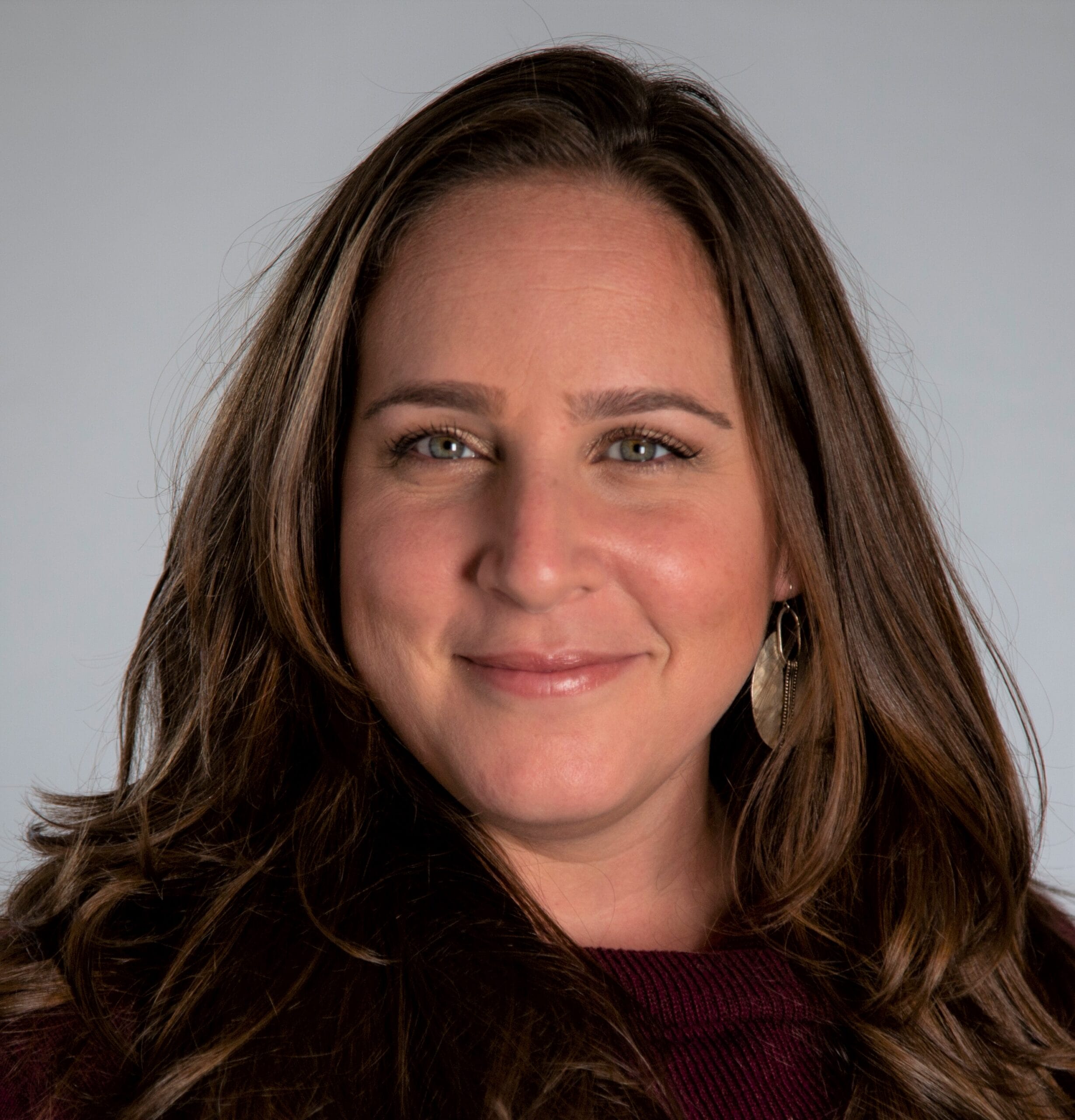 Woman with long brown hair, wearing a maroon top and earrings, smiling in front of a gray background.