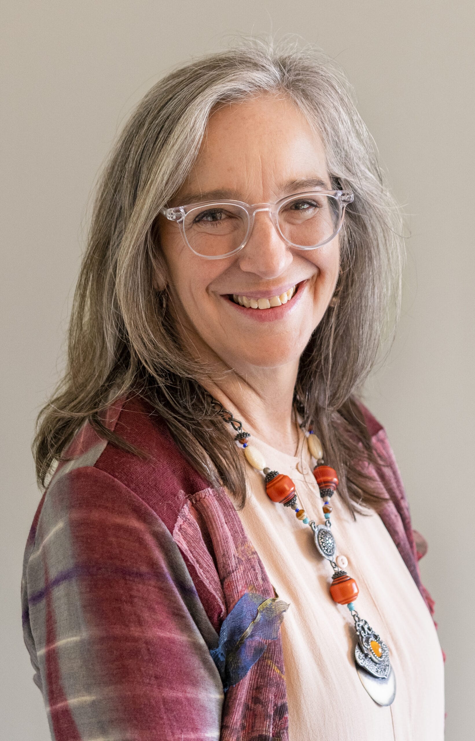 Smiling woman with gray hair, glasses, and a colorful necklace, wearing a red plaid jacket.