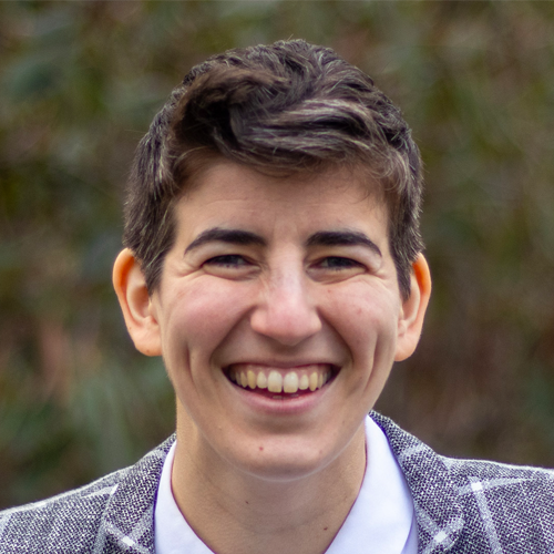Person with short dark hair and a gray blazer smiling outdoors with a blurred green background.
