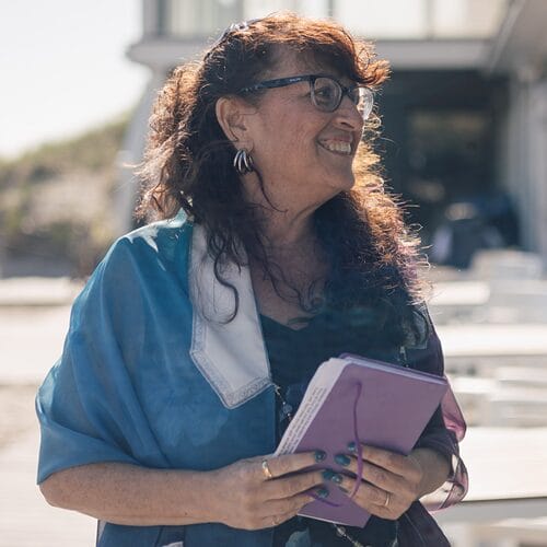 Smiling woman with glasses holds a purple notebook outdoors, wearing a blue shawl and earrings.