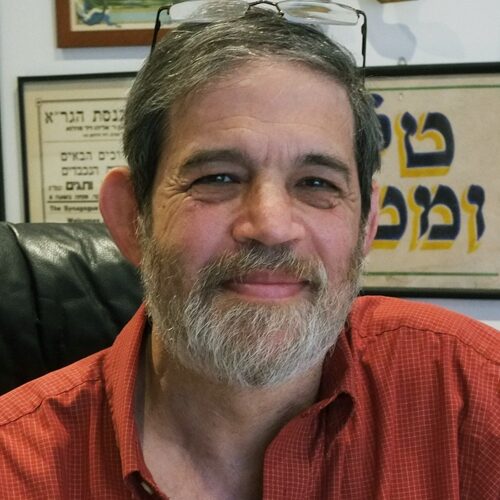 A man with gray hair and beard, wearing glasses and a red shirt, smiles while sitting indoors.