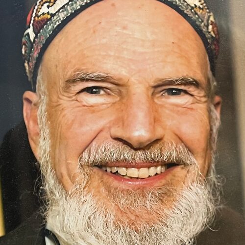 Elderly man with a white beard, smiling and wearing a colorful patterned hat.