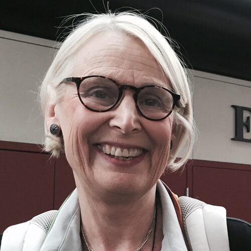 Older woman with short gray hair, glasses, and a light jacket smiling at the camera.