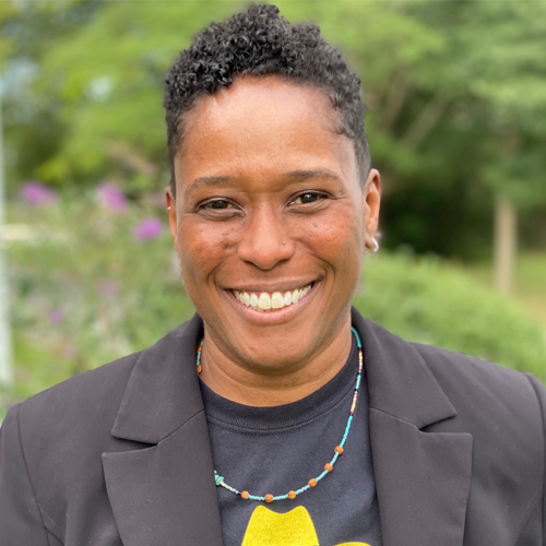 Person with short curly hair in a black blazer smiling outdoors with greenery in the background.