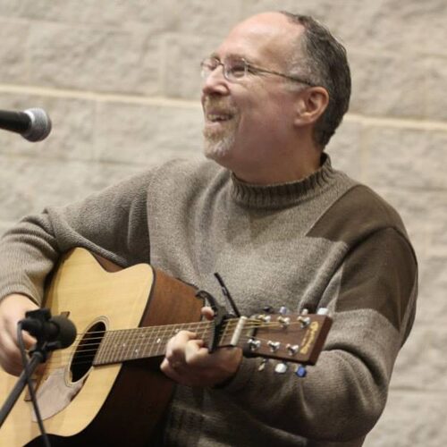 A smiling man plays an acoustic guitar and sings into a microphone, wearing glasses and a brown sweater.