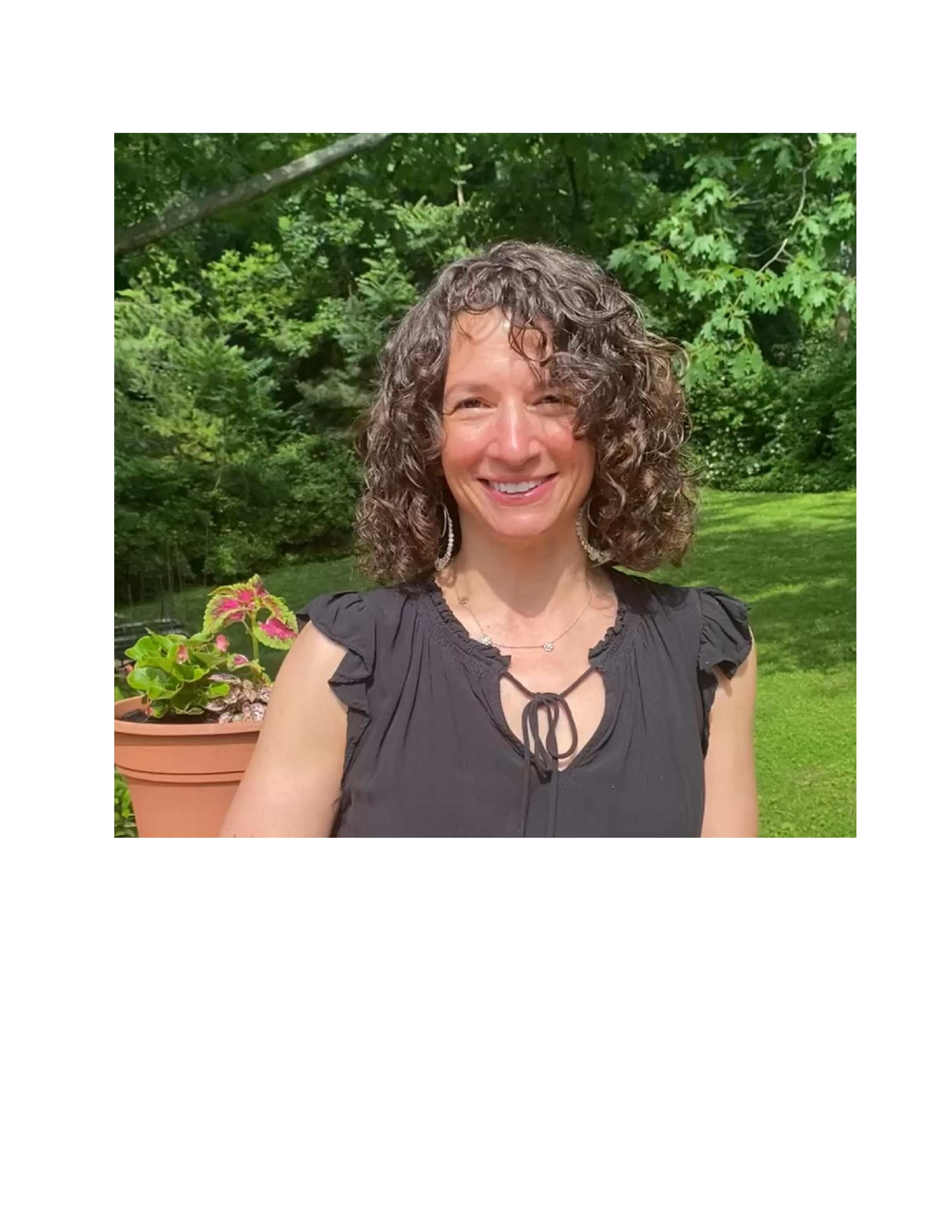 Woman with curly hair smiling outdoors, standing in front of greenery and a potted plant.