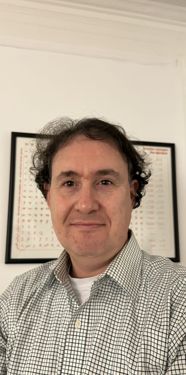 Man with dark curly hair in a checkered shirt smiling, with framed charts on the wall behind him.