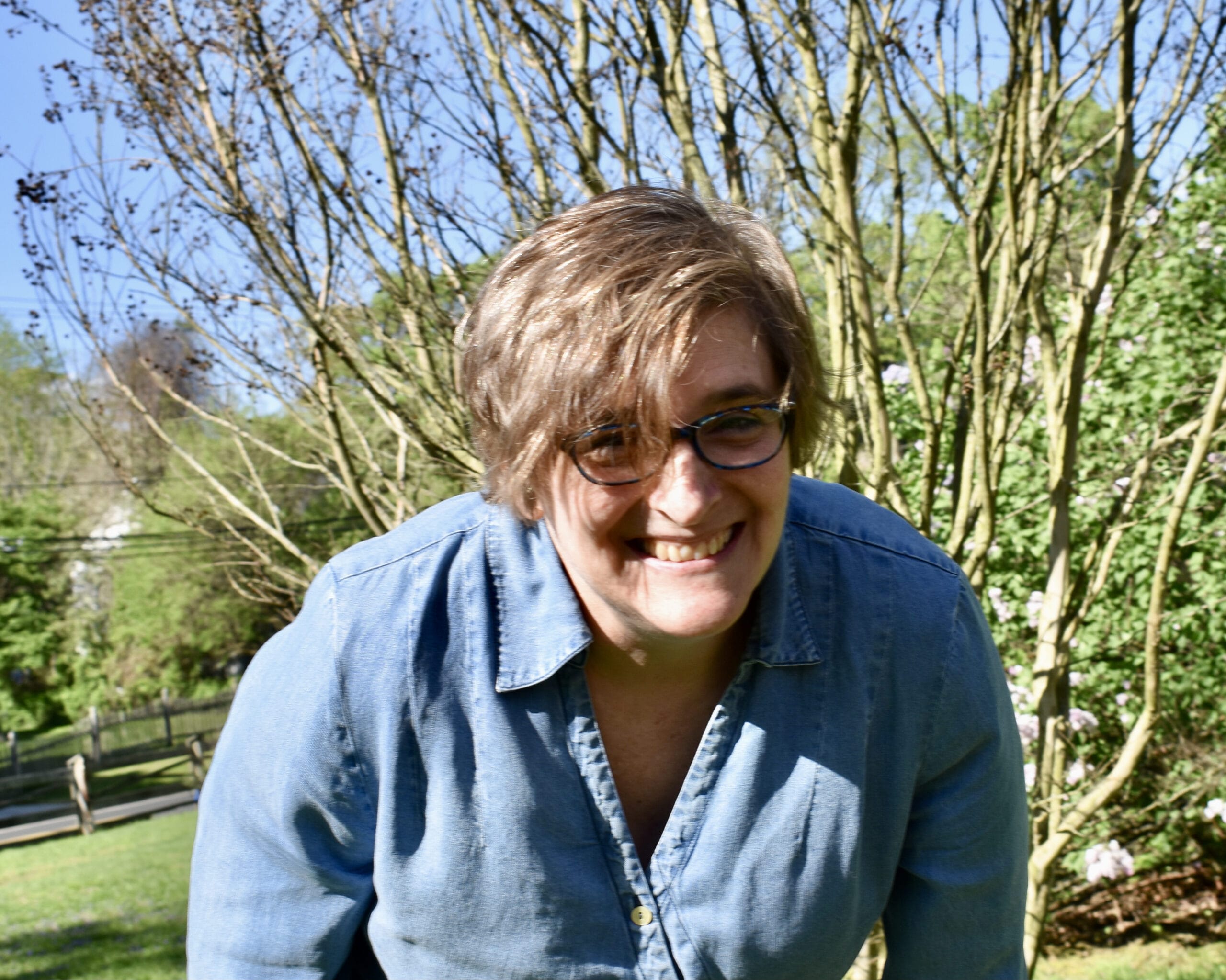 Smiling person with short hair and glasses wearing a denim shirt, standing outdoors by trees on a sunny day.