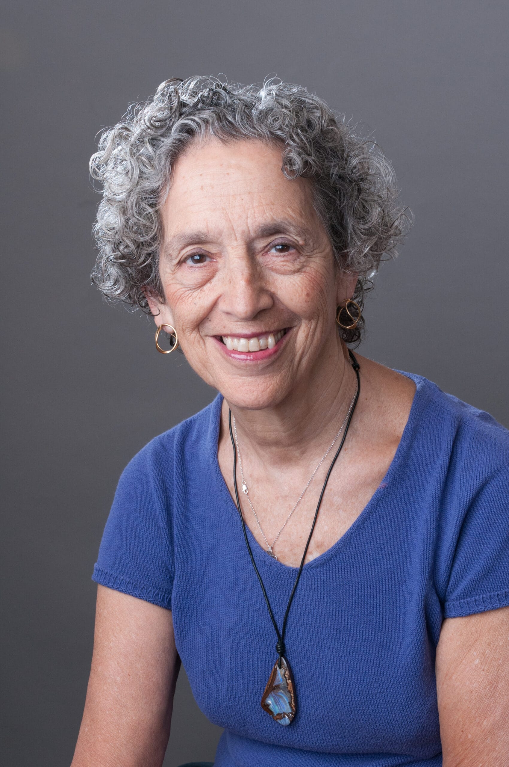 Smiling older woman with curly gray hair, wearing a blue top and a pendant necklace, on a gray background.