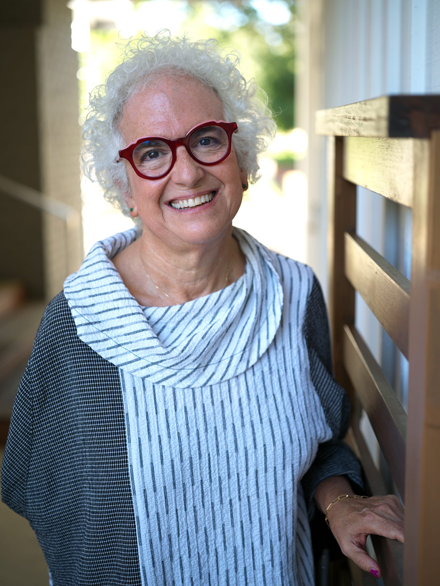 Smiling older woman with curly white hair and red glasses, wearing a striped top, standing outdoors.