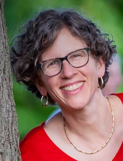 Woman with short curly hair, glasses, and gold jewelry smiling outdoors, wearing a red top.