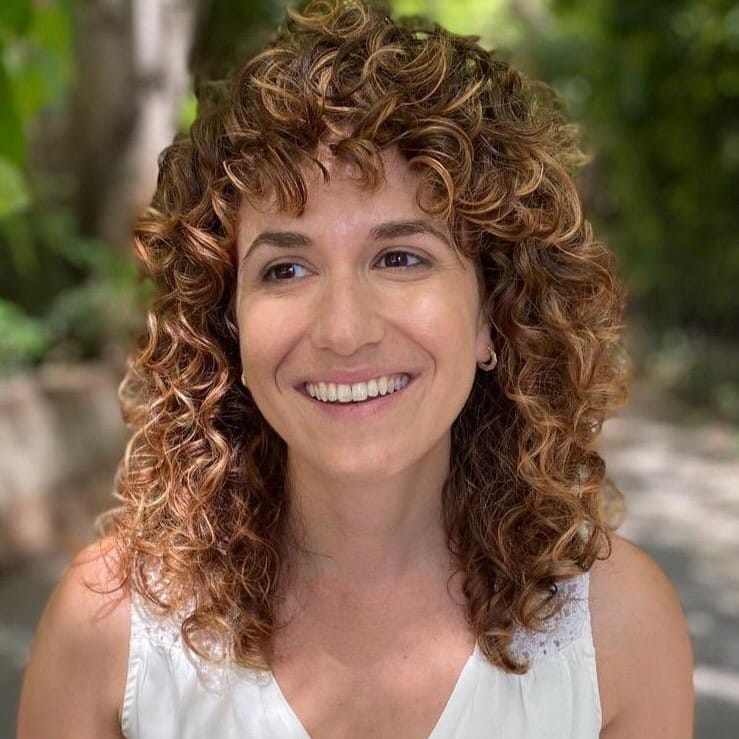 A woman with curly hair and a white top smiles outdoors with greenery in the background.