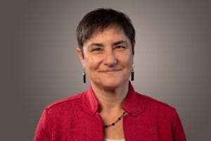 Smiling person with short dark hair, wearing a red jacket, earrings, and a necklace, against a plain background.
