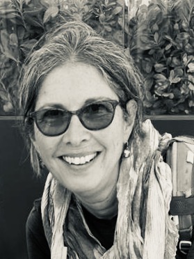 Smiling woman with glasses and scarf outdoors, in front of leafy green plants.