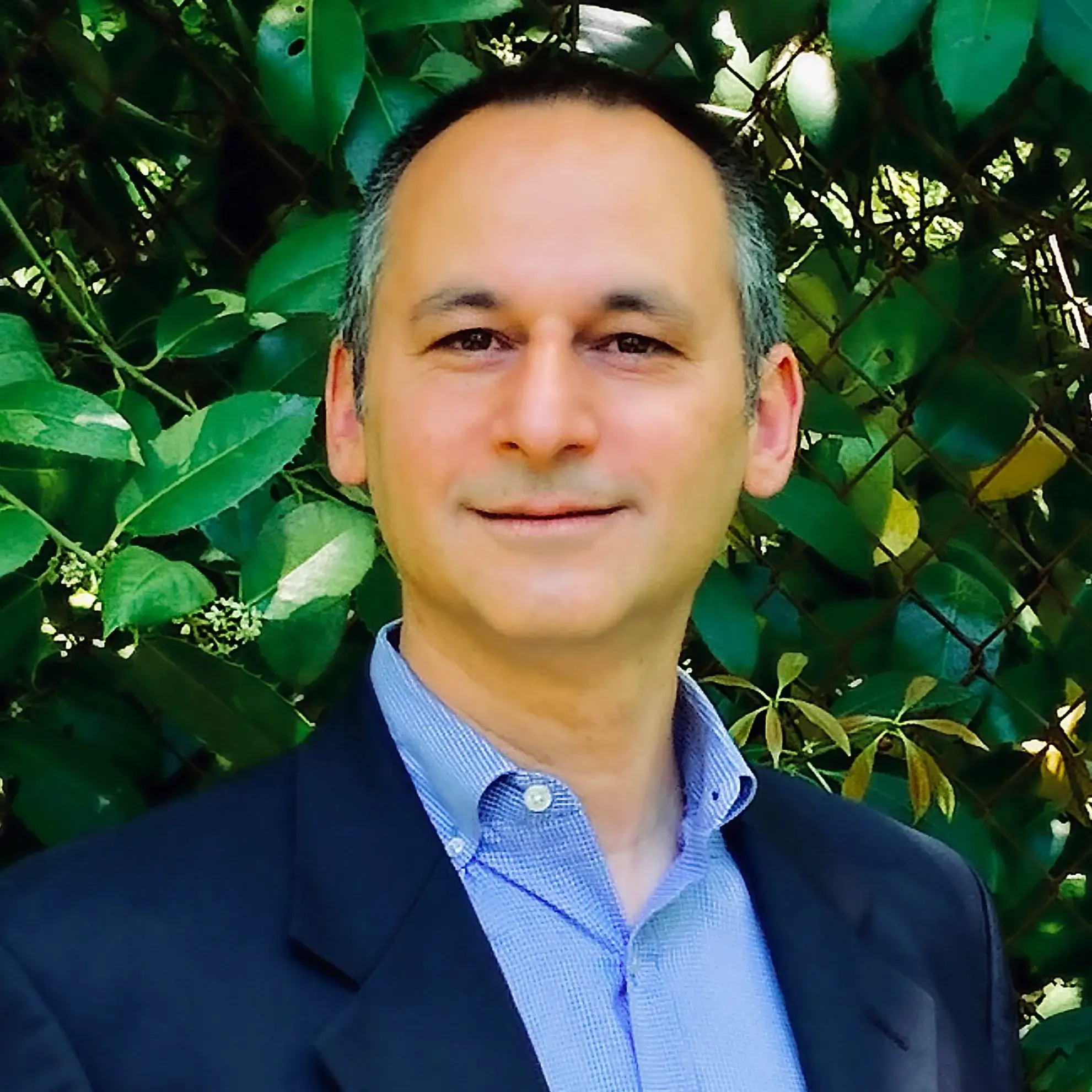 A man in a suit smiles in front of green leafy plants outdoors.