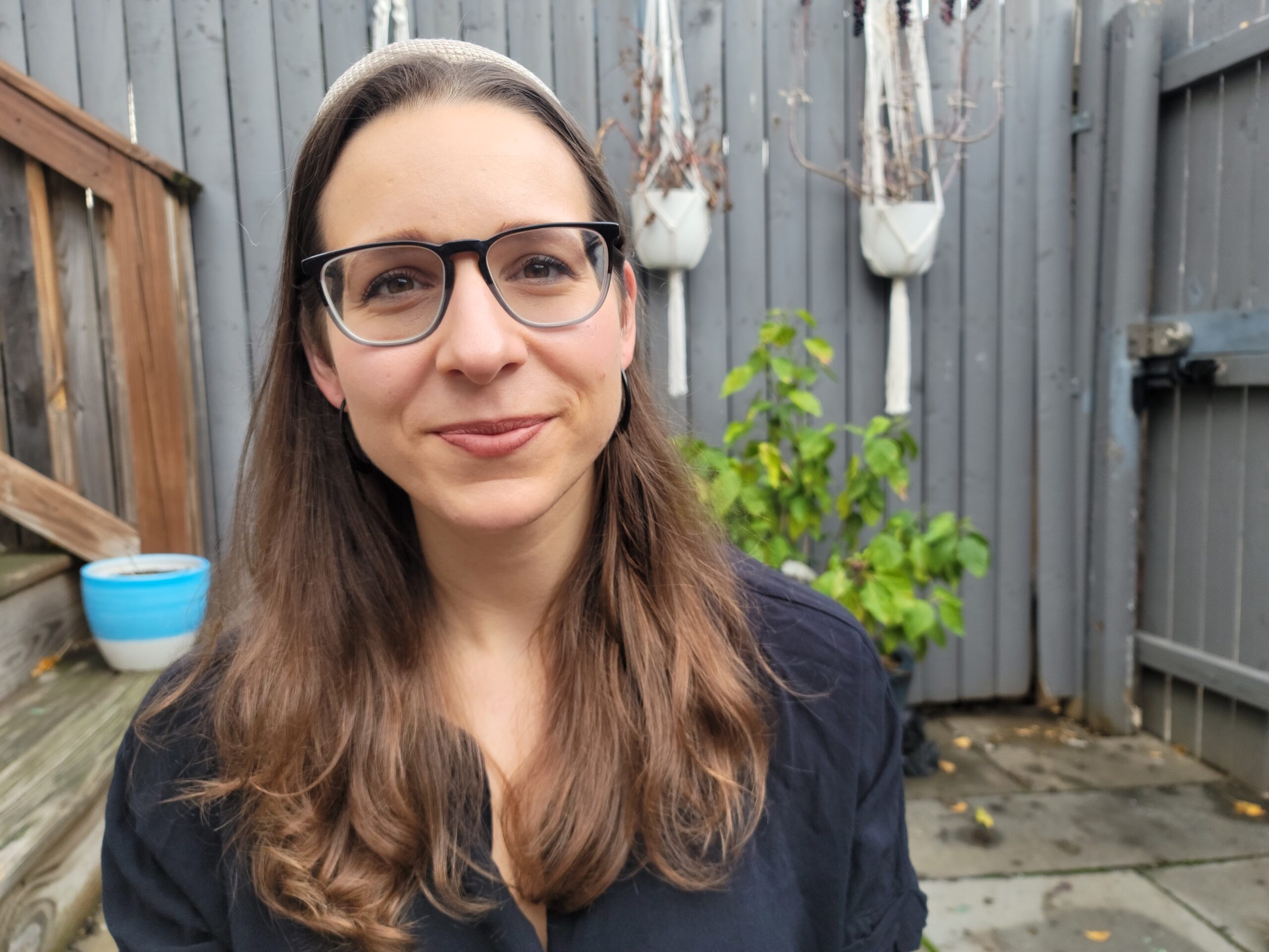 A woman with long brown hair and glasses smiles outdoors in a fenced yard with plants.