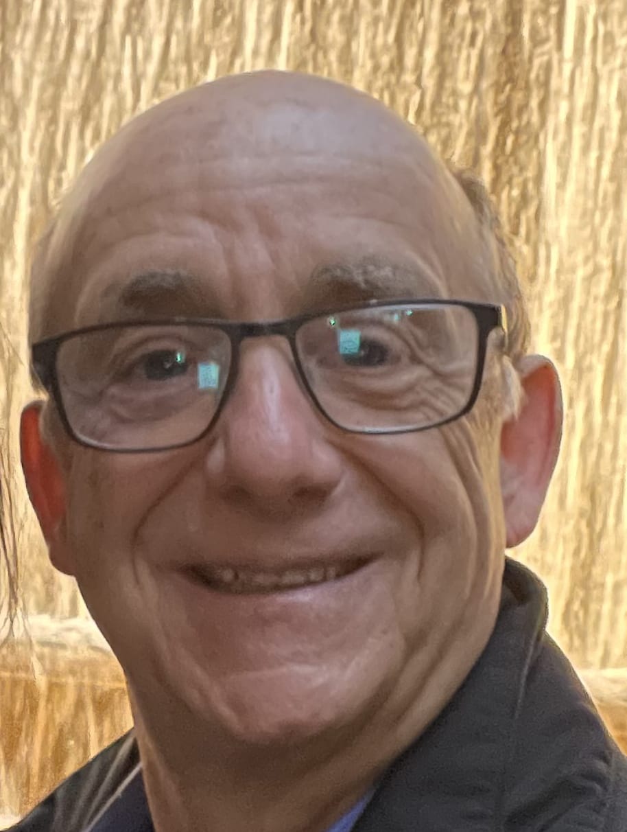 Smiling older man with glasses, standing in front of a golden, cascading water background.